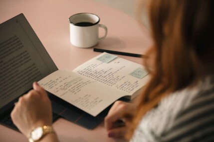 A person’s hands resting on a laptop keyboard and notebook at a pink desk with a cup of coffee.