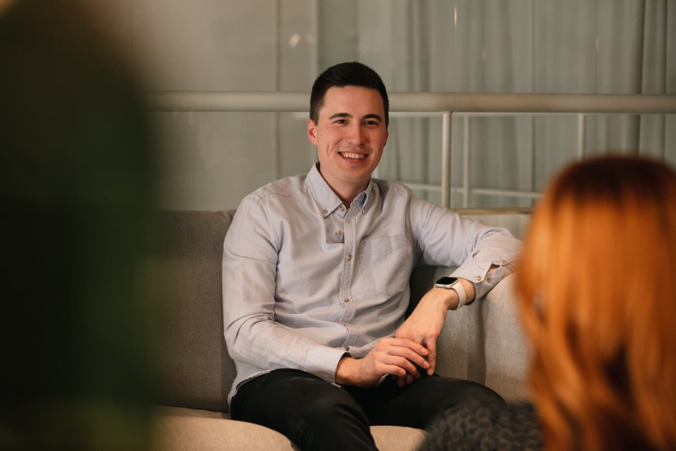 A man sitting on a gray couch in a living room with a plant in the background.